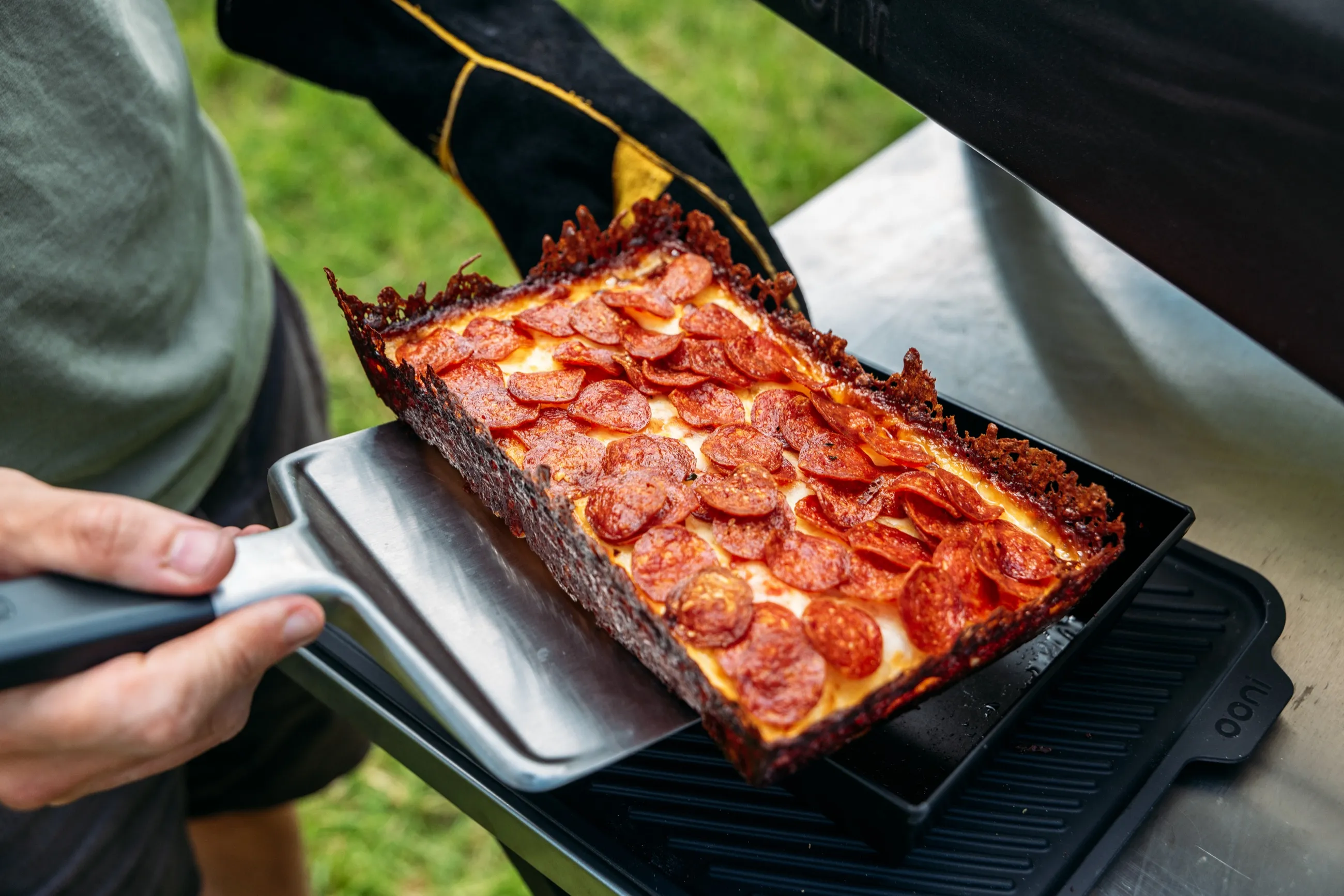 Detroit-style pepperoni pizza being lifted from an outdoor pizza oven